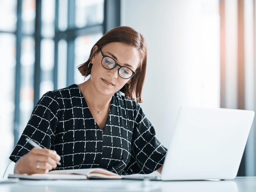 Woman writing on a note pad in front of a laptop
